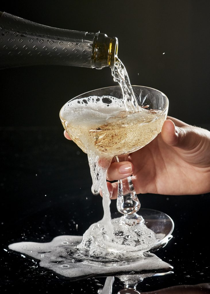 A bottle of champagne pouring into a glass against a dark background. The glass is overflowing. 