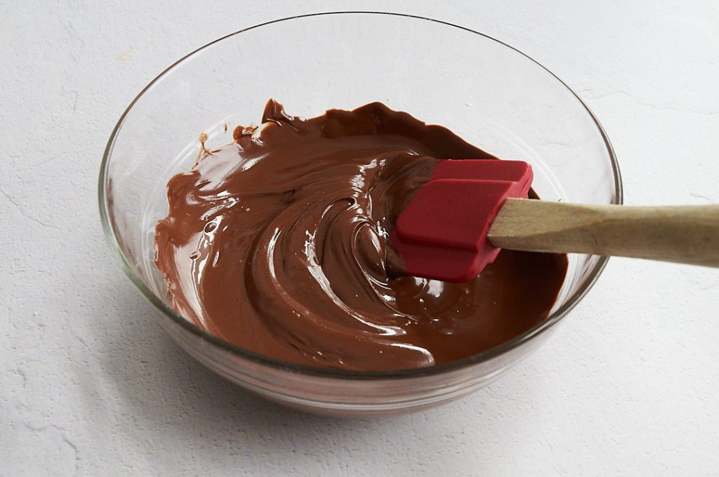 A glass bowl full of melted chocolate, being stirred by a red silicone spatula with a wooden handle. 