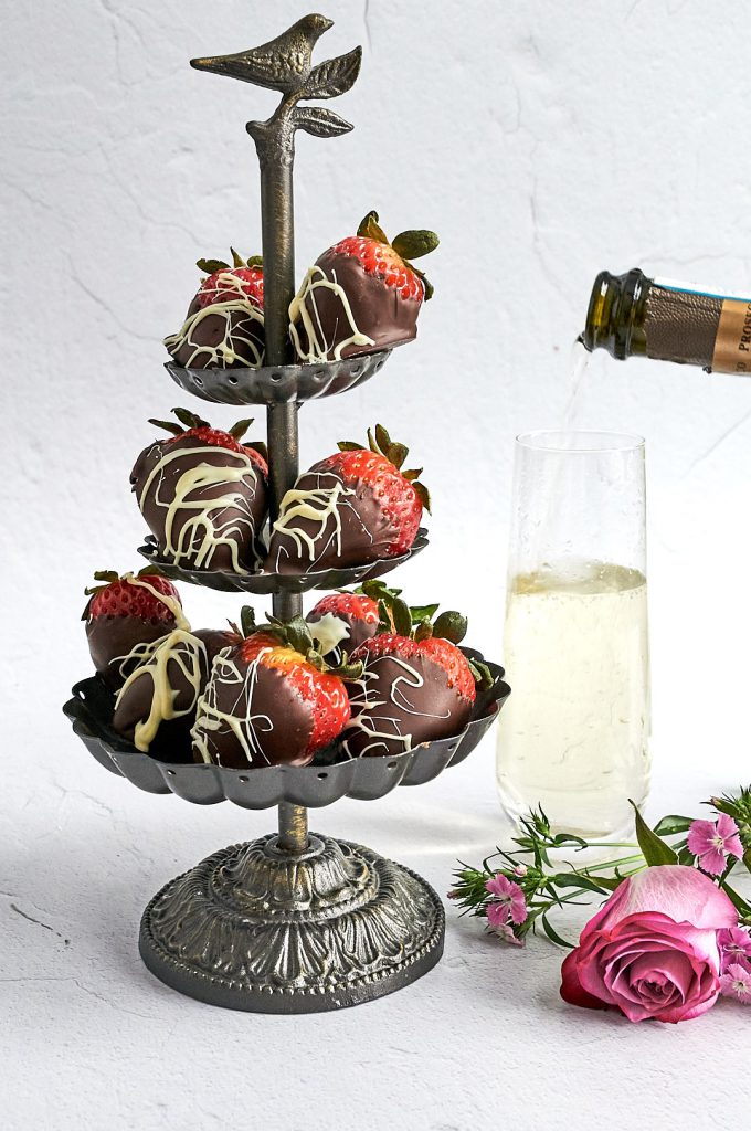 A metal display holding chocolate dipped strawberries. On the right are pink roses and a glass of champagne.