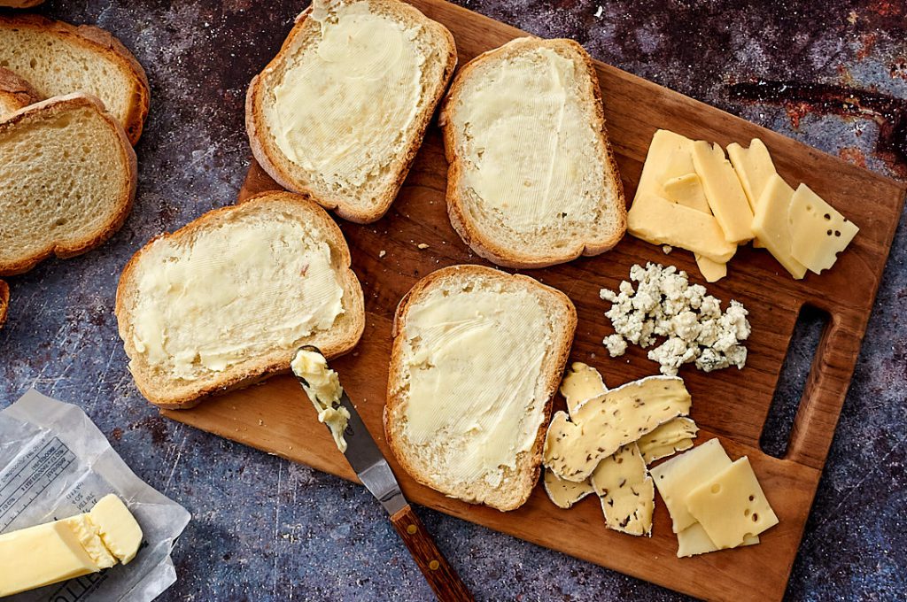 A wooden cutting board with four slices of quality bread slathered in butter and different types of cheese. 