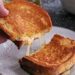 A grilled cheese sandwich sliced in half. A hand holds up half of the sandwich and strings of melted cheese are visible.