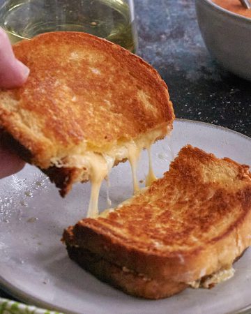 A grilled cheese sandwich sliced in half. A hand holds up half of the sandwich and strings of melted cheese are visible.