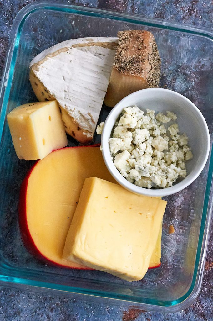 A glass storage container full of assorted cheeses, including Gouda, brie, blue, Gruyere and Havarti. 