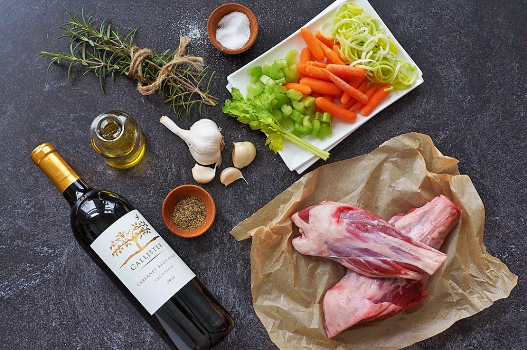 Lamb shank, garlic, olive oil, rosemary, wine, and a white plate with carrot, celery, and onion on a dark countertop.