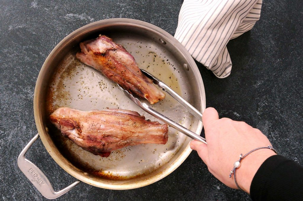 Two lamb shanks searing in an All-Clad pan. A woman is using metal tongs on one of the lamb shanks. 