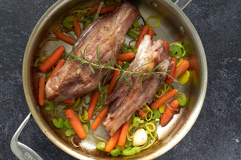 Two lamb shanks in an All-Clad pan with carrot, celery, onion, and a spring of rosemary on top. 