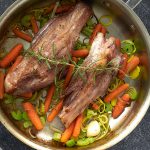 Two lamb shanks in an All-Clad pan with carrot, celery, onion, and a spring of rosemary on top.