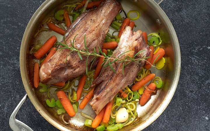 Two lamb shanks in an All-Clad pan with carrot, celery, onion, and a spring of rosemary on top.