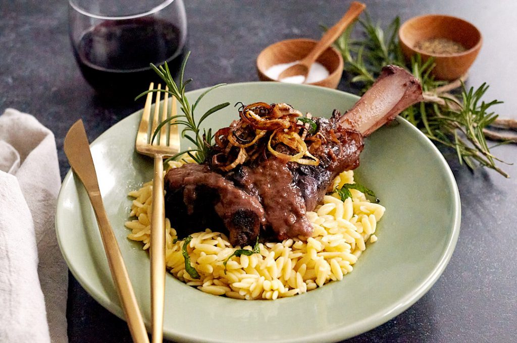 A green plate with a braised lamb shank on minted orzo, topped with fried leek and rosemary, accompanied by a glass of wine.